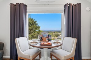 Ein Essbereich mit Tisch und zwei Stühlen, ein Fenster mit Vorhängen und Blick ins Grüne.
