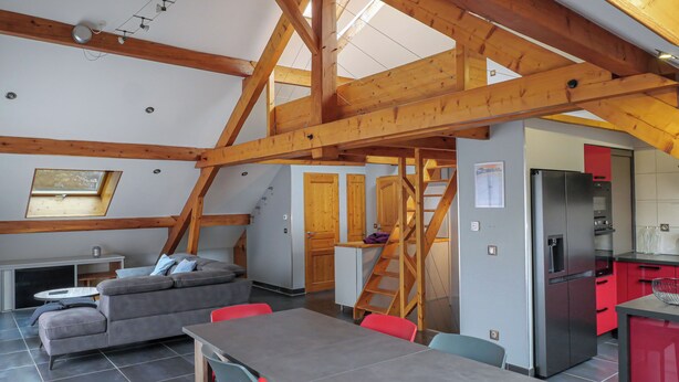 A loft-style room with exposed wooden beams, a sofa, a table, and a kitchen area with a refrigerator and red cabinets.