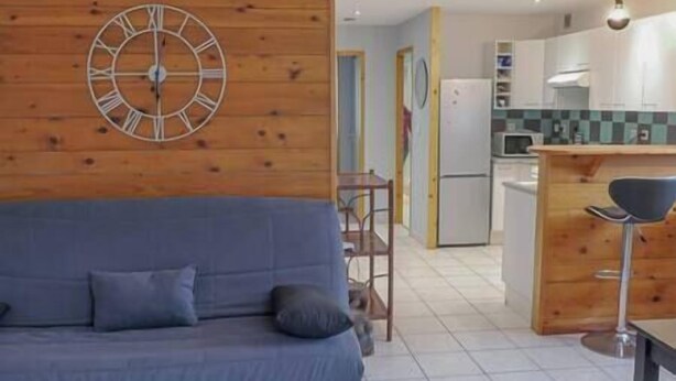 Una sala moderna con un sofá azul, un reloj de pared de madera y una zona de cocina con armarios blancos y un microondas.