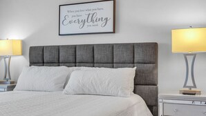 A neatly made bed with a grey upholstered headboard, two bedside lamps, and a framed inspirational quote on the wall.