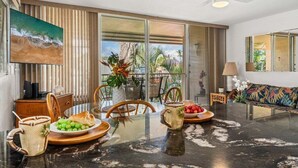 Un área de comedor moderna con una mesa de vidrio, sillas y un sofá. Hay un televisor en la pared y un ventilador de techo.