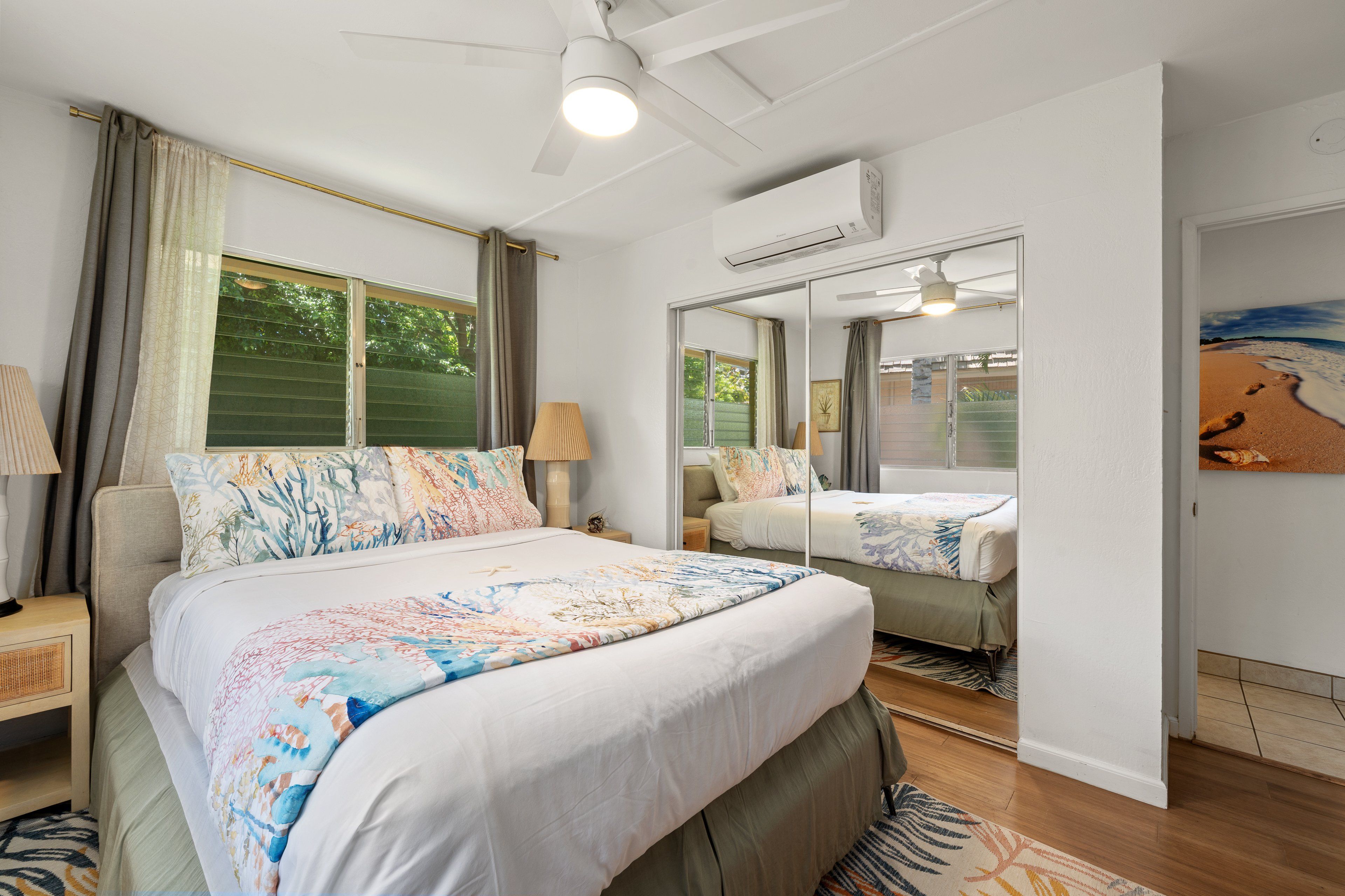 Een slaapkamer met een groot bed, een plafondventilator, een raam met gordijnen en een spiegel die de kamer weerspiegelt.