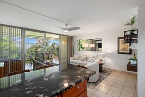 A modern living room with a glass dining area, a sofa with floral cushions, a ceiling fan, and a view of the outdoors.
