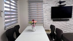 A modern dining area with a white table, black chairs, a vase with flowers, and a flat-screen TV mounted on a textured wall. - American Standard/2Bedroom House (Cagayan de Oro)