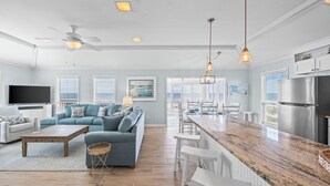 A modern living room with a sofa, coffee table, and TV, a kitchen with a refrigerator and island, and a dining area with chairs and a table.