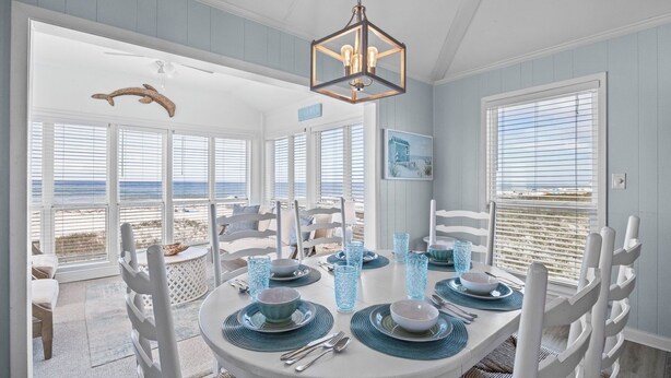 A dining area with a view of the beach, white furniture, and a hanging light fixture.