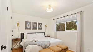 Un dormitorio ordenado con cama, mesitas de noche, un banco y una ventana con cortinas.