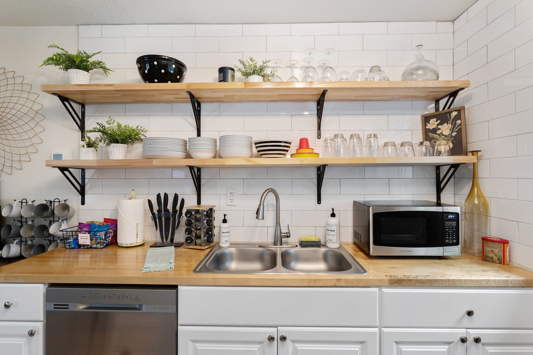 Una cocina con armarios blancos, un lavamanos de acero inoxidable, un microondas y estantes con vajilla y plantas.