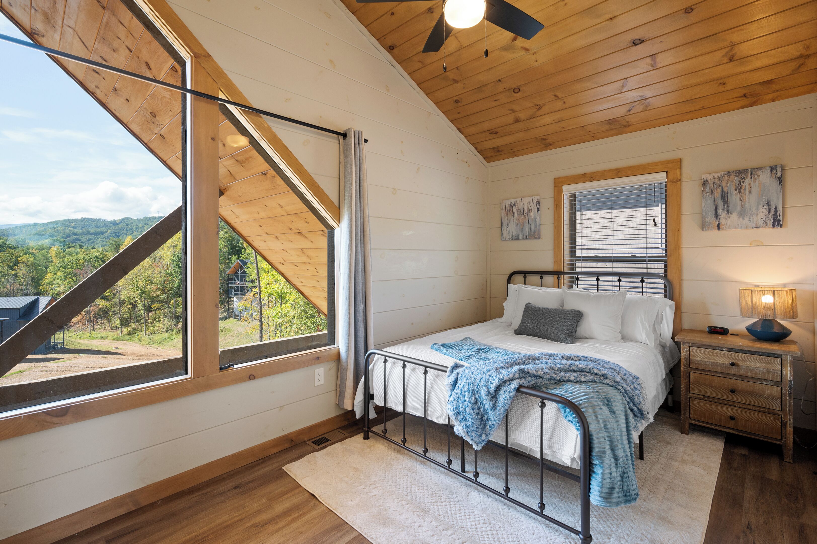 A bedroom with a bed, a nightstand, a window with a view of the outdoors, and a ceiling fan.