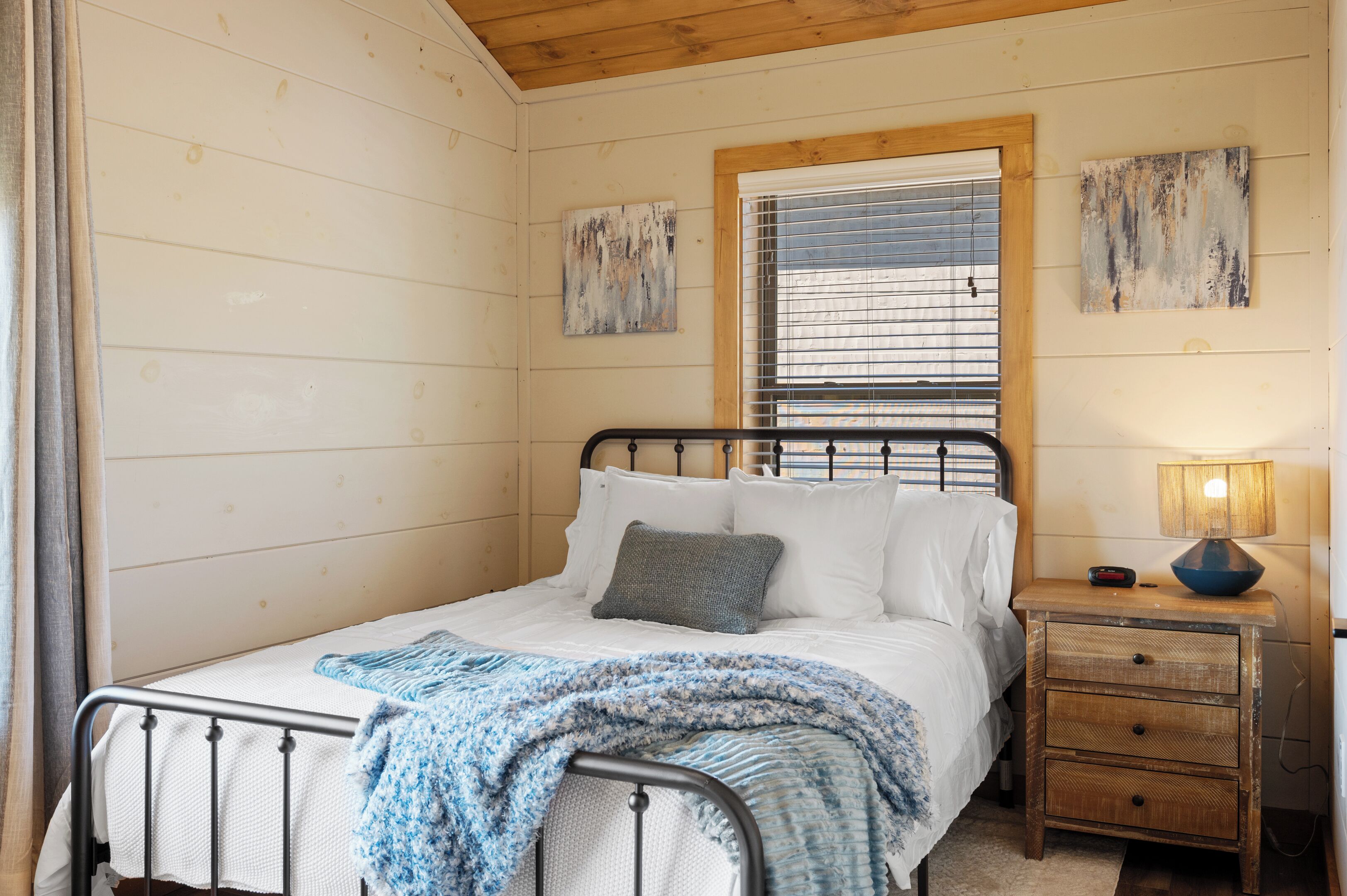 A bedroom with a metal bed frame, white bedding, a blue and white blanket, a wooden nightstand with a lamp, and two abstract paintings on the wall.