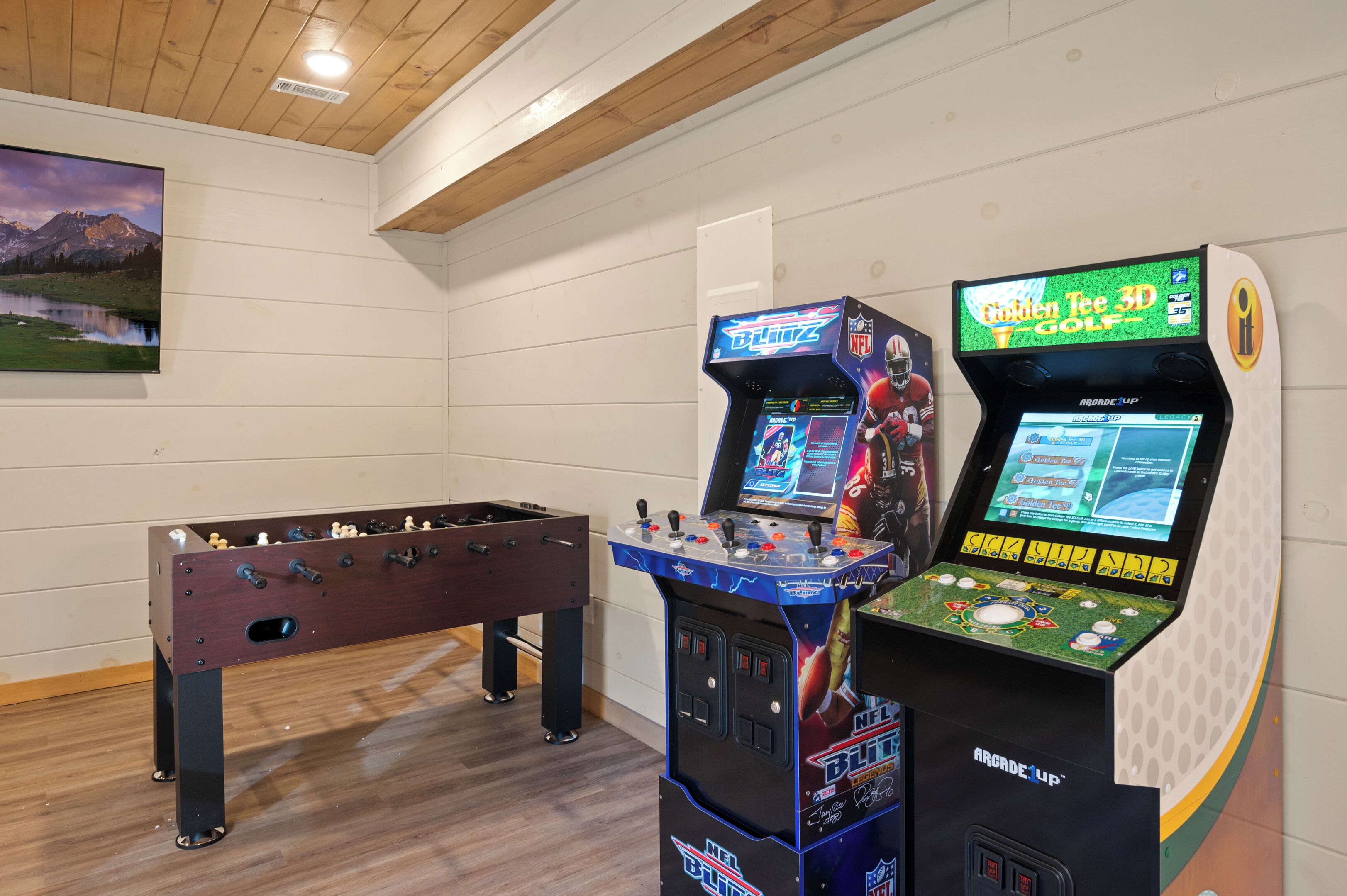 A room with a foosball table, two arcade machines, and a painting of a mountain landscape.