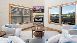 A cozy living room with a fireplace, comfortable seating, and large windows offering scenic views. - New cabin! Great Location! 5 Min to Parkway! Private pool, hot tub, & game room! (Pigeon Forge)