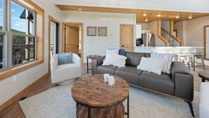 A modern living room with a grey sofa, white armchair, and a round wooden coffee table. There is a kitchen area with a sink and a staircase leading to another level. - New cabin! Great Location! 5 Min to Parkway! Private pool, hot tub, & game room! (Pigeon Forge)