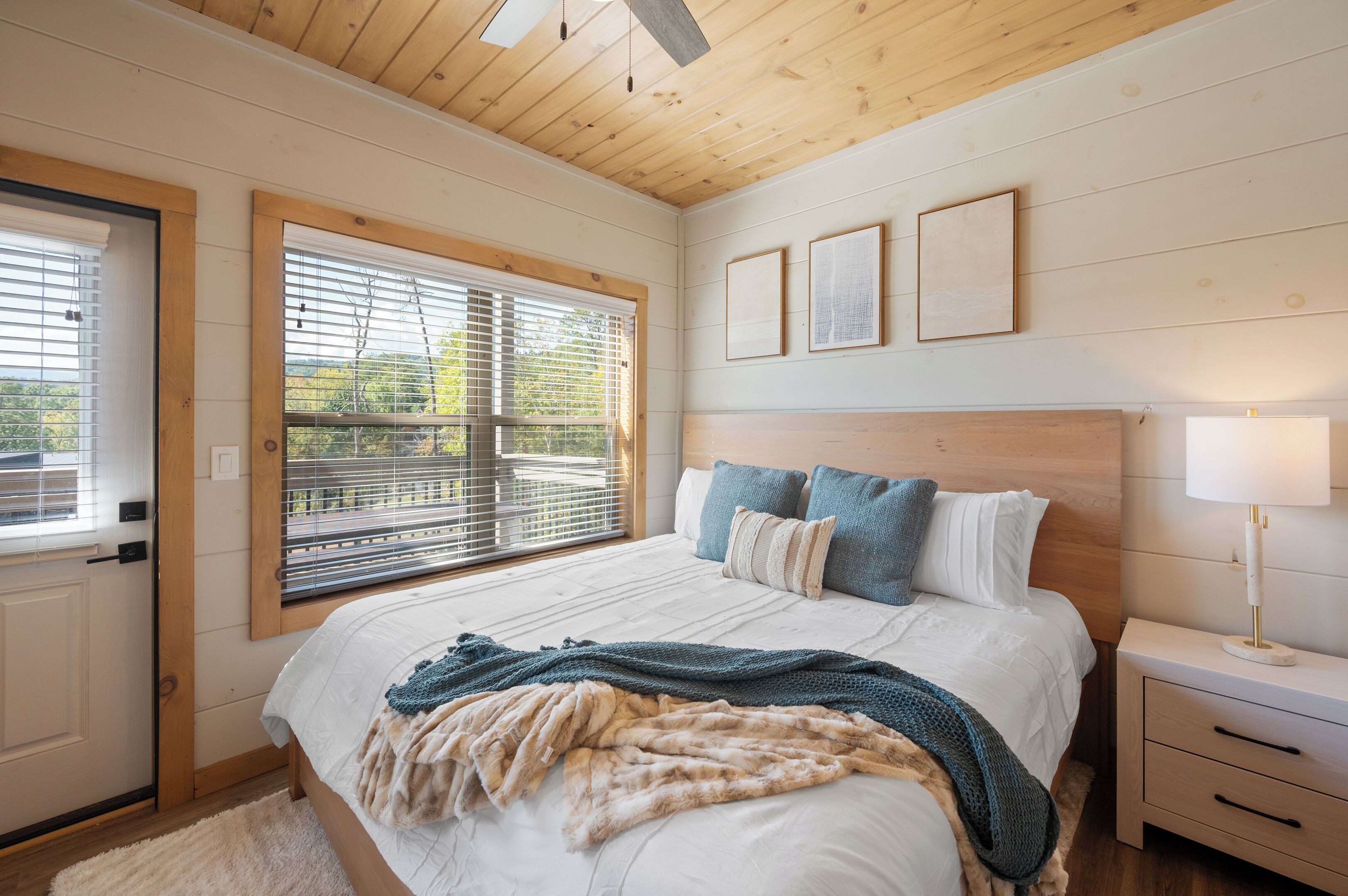 Une chambre avec un plafond en bois, un grand lit avec une literie blanche et bleue, une table de chevet avec une lampe et une fenêtre avec des stores.