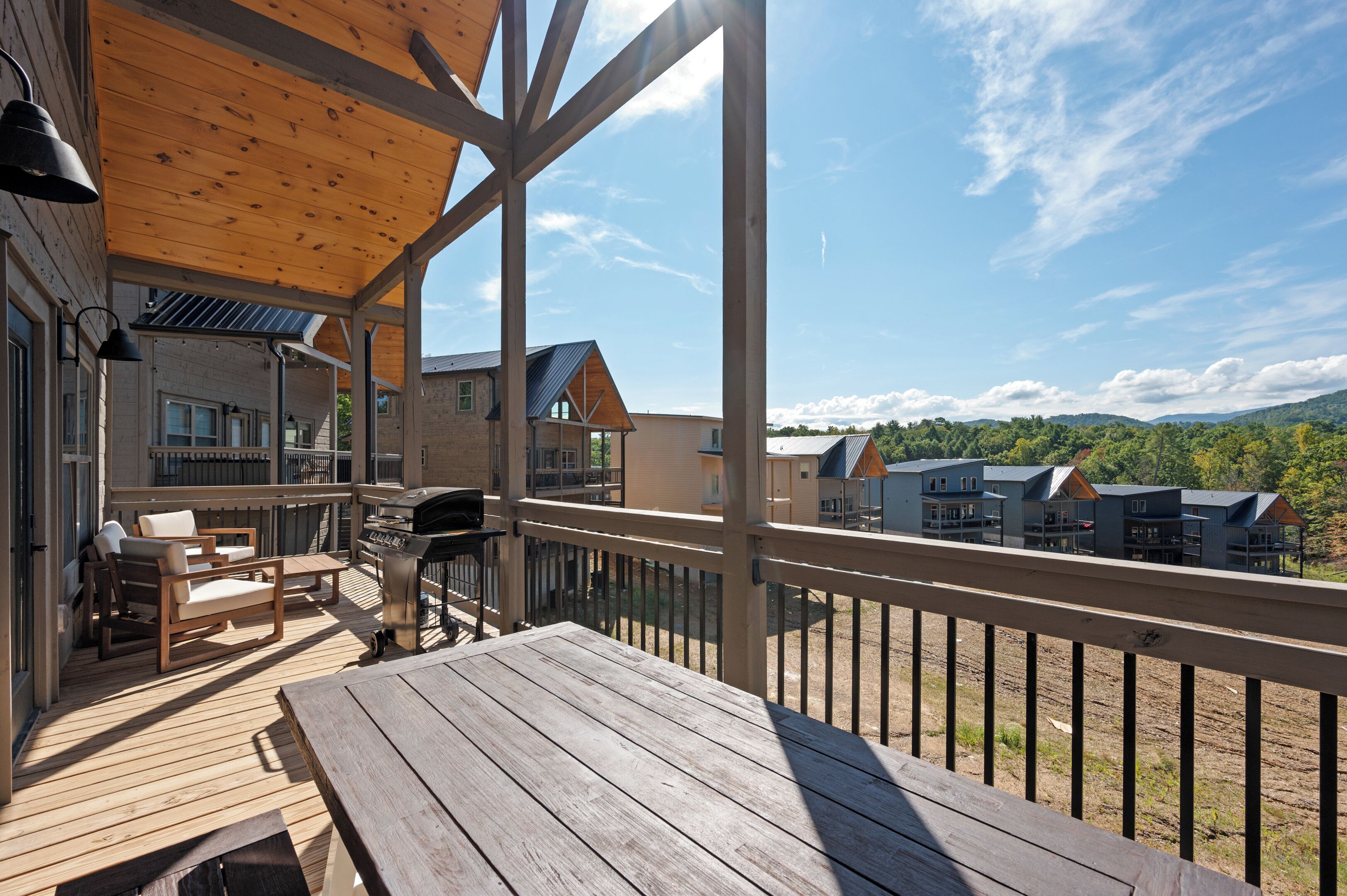 Una terraza de madera con mobiliario exterior, una parrilla de barbacoa y vistas de edificios y colinas circundantes.
