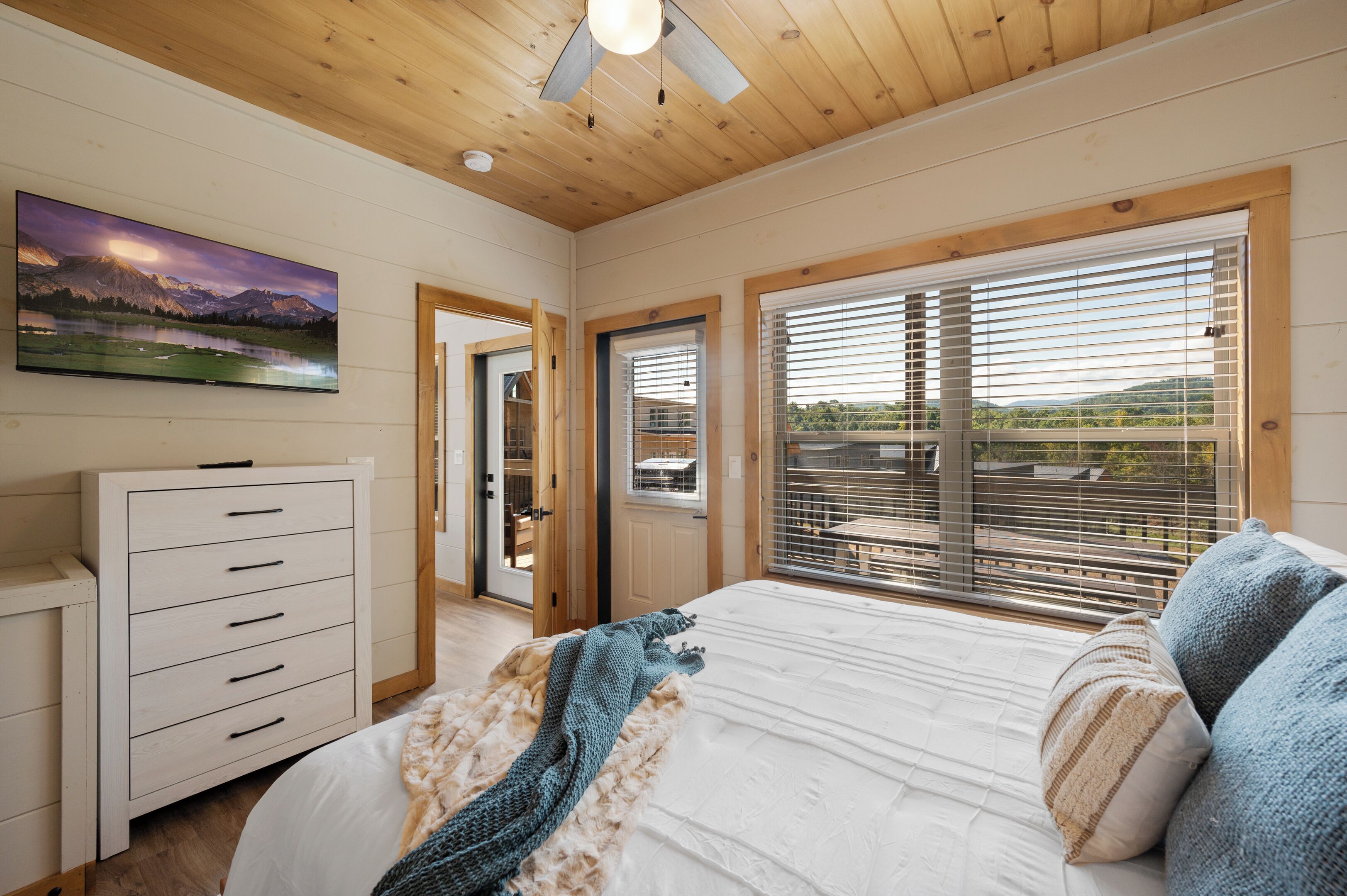 A bedroom with a bed, a dresser, a ceiling fan, a large window with blinds, and a painting on the wall.