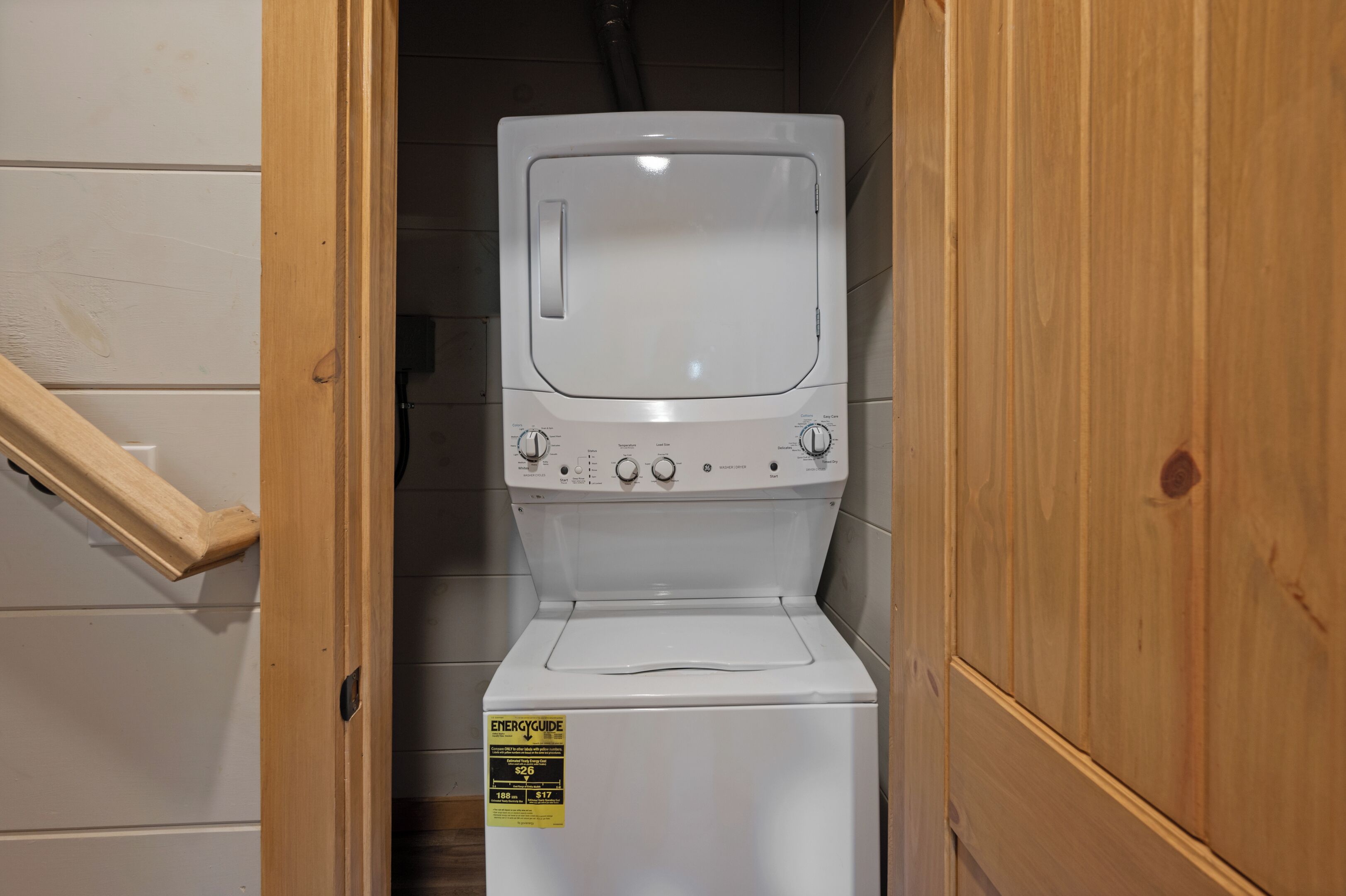 A compact washer and dryer unit in a narrow hallway with wooden walls and a wooden staircase.