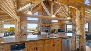 A log cabin interior with a kitchen featuring granite countertops, wooden cabinets, and a stone wall. - POOL in Huge Cabin, Sleeps 43+, Tons of Parking (Fish Haven)