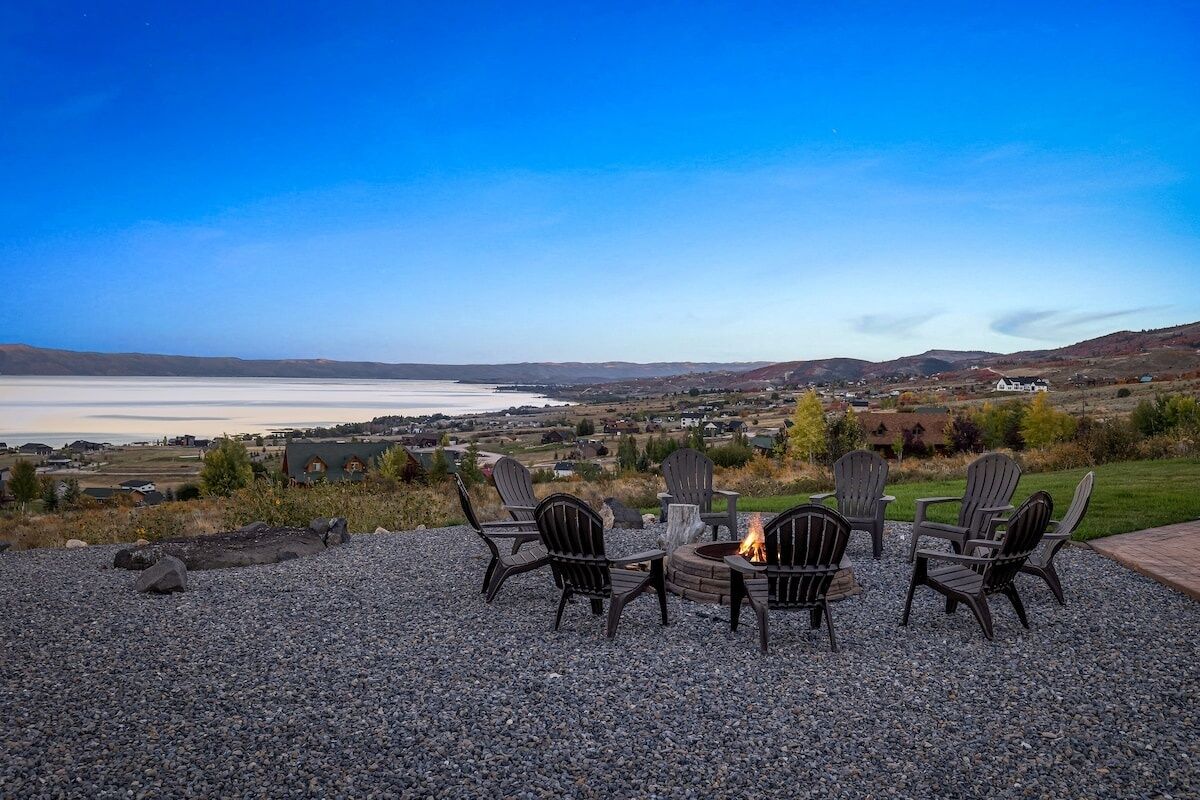 Un patio de grava con sillas Adirondack y un pozo de fuego con vistas a un lago y una ciudad distante.