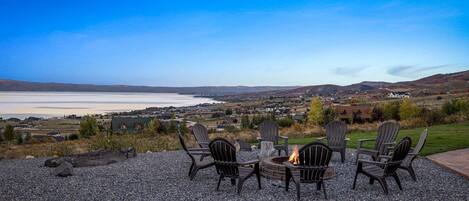 Un patio de grava con sillas Adirondack y un pozo de fuego con vistas a un lago y una ciudad distante.