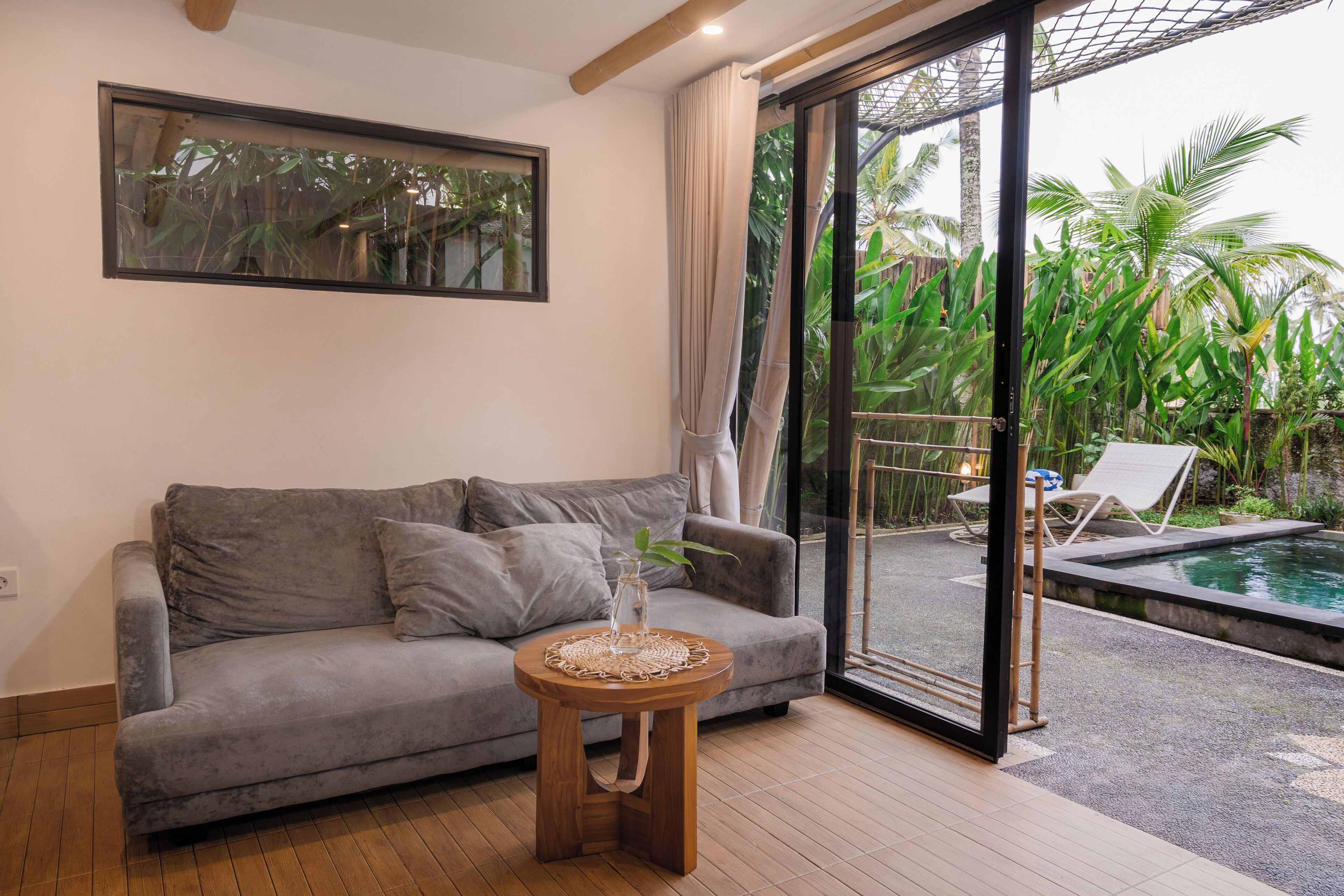 Une pièce chaleureuse avec un canapé gris, une petite table en bois surmontée d’un vase et une porte vitrée donnant sur un espace piscine équipé de chaises longues.