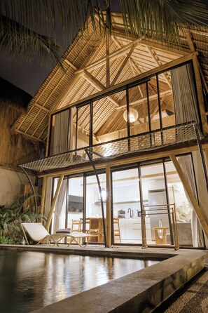 A two-story building with a thatched roof, large windows, and a pool area with lounge chairs. - Aranya Villa by Betterplace (Ubud)