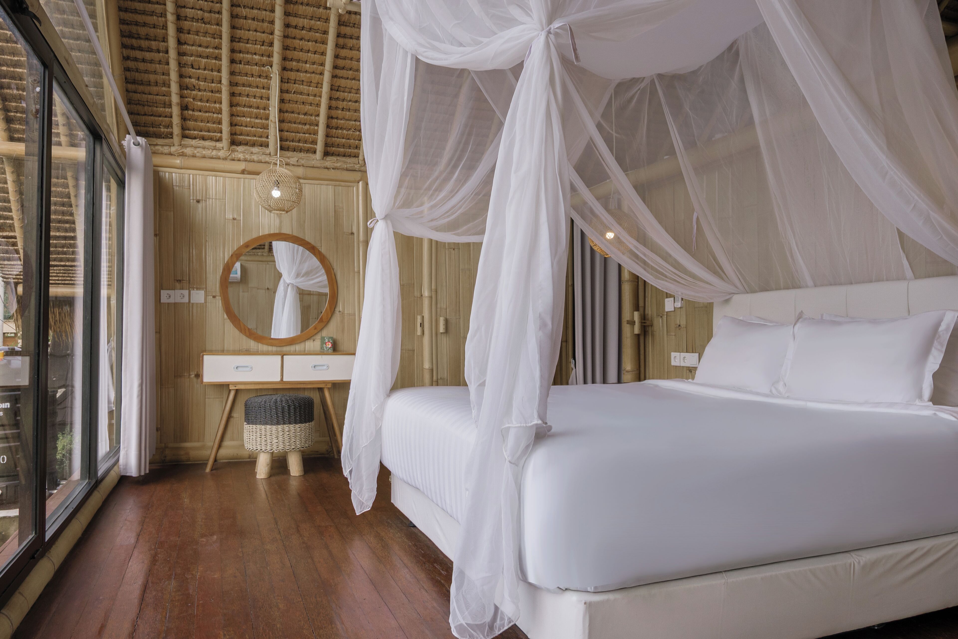 Een slaapkamer met een hemelbed, een ronde spiegel, een houten dressoir en een rieten kruk.
