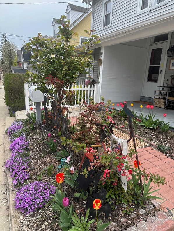 En trädgård vid ett bostadshus med ett stort utbud av blommor, ett litet träd och en fågelskulptur.
