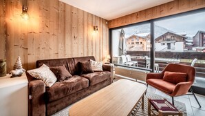 Uma sala de estar moderna com parede de madeira, um sofá marrom, uma poltrona vermelha e uma grande janela com vista para uma aldeia montanhosa.