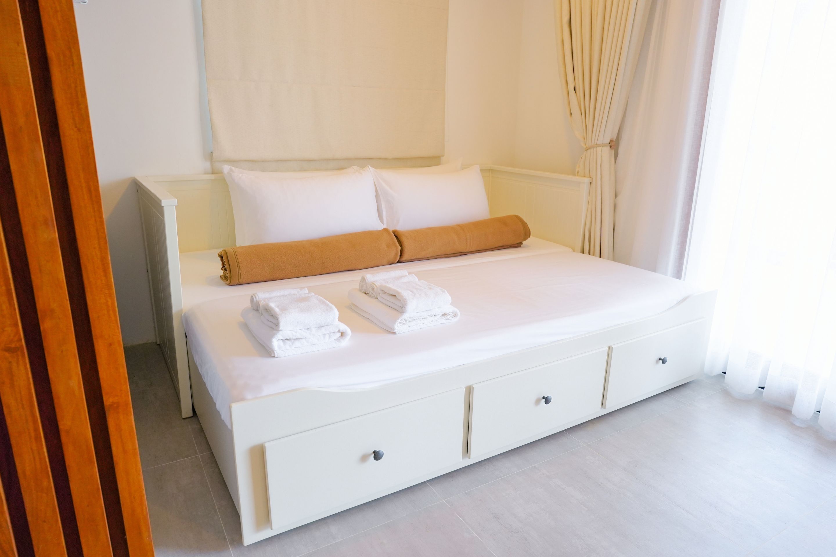 A neatly made bed with white linens and a brown towel, a white bedside table with drawers, and a window with sheer curtains.