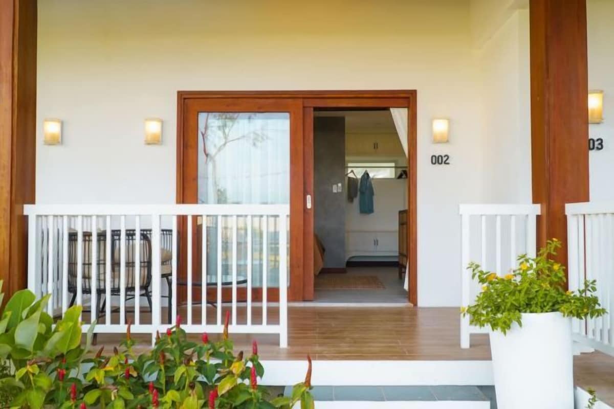 A hotel entrance with a wooden door, numbered rooms, and a small outdoor seating area with chairs.