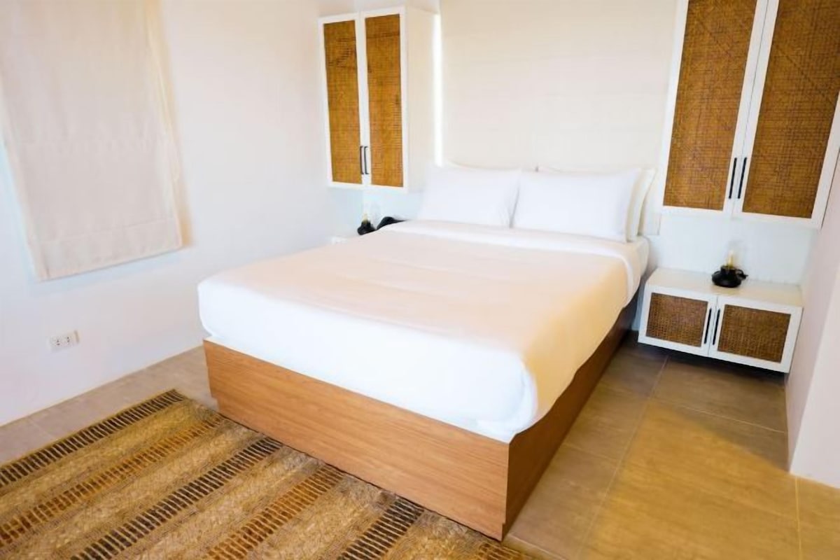 Una habitación de hotel moderna con una cama grande, un cabecero de madera, una mesita de noche pequeña y un armario con puerta trenzada.