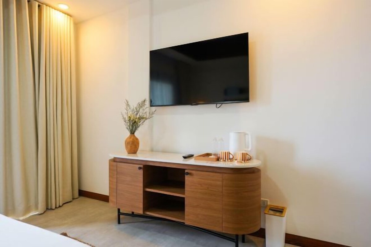 A hotel room with a wooden cabinet, a flat-screen TV mounted on the wall, a vase with dried plants, and a trash bin.