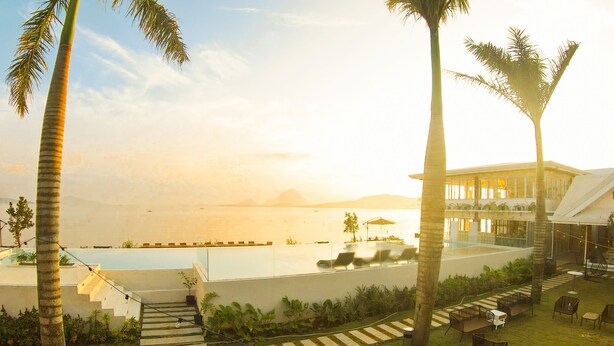 A resort pool area with palm trees, lounge chairs, and a building with large windows overlooking the pool. - Caza Guesthouse Iyang (Concepcion)
