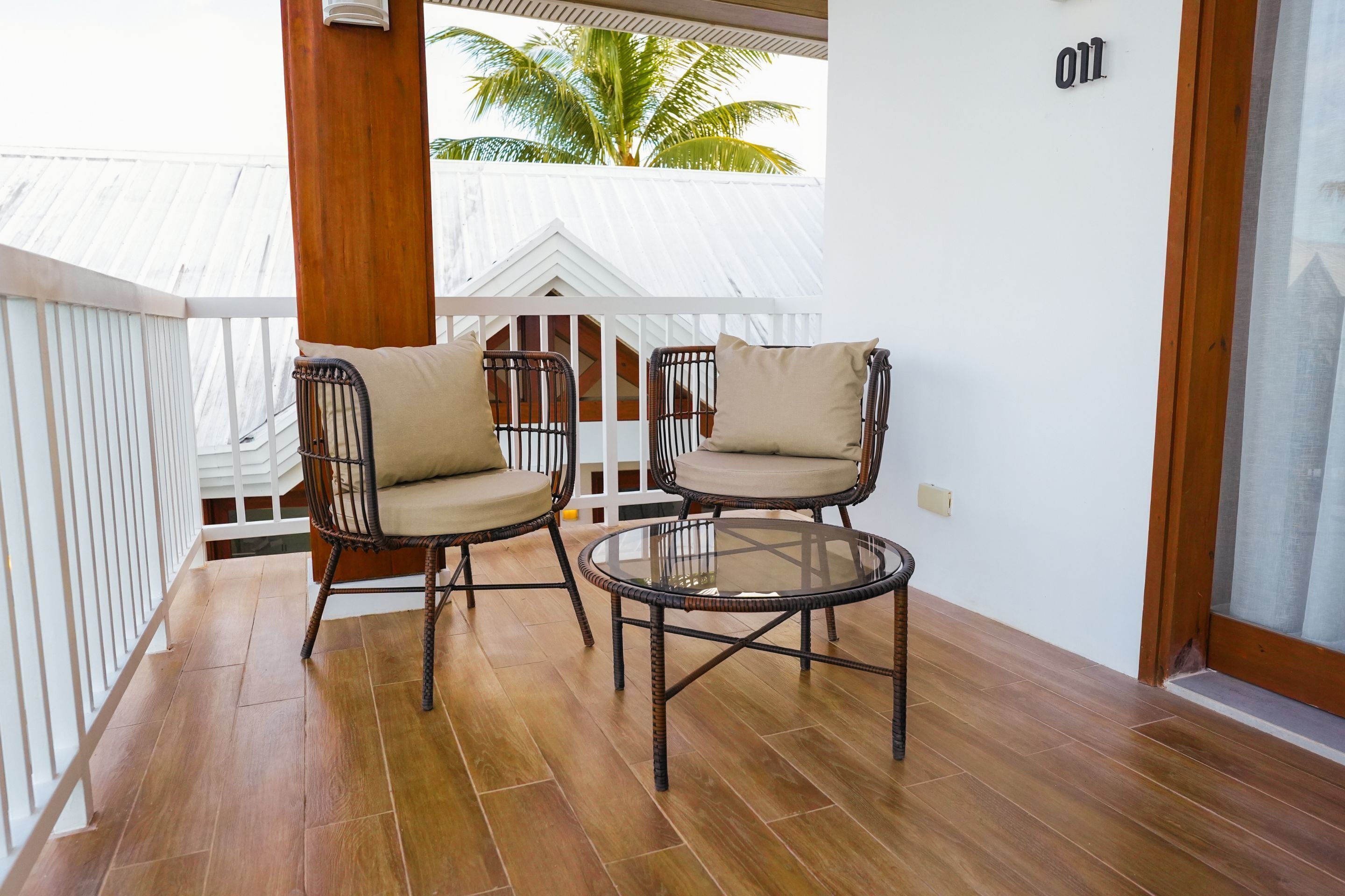 Een balkon met twee rieten stoelen en een glazen tafel, wit hekwerk en uitzicht op een palmbomen.
