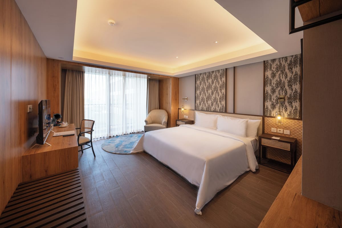 A modern hotel room with a large bed, a desk with a chair, a TV, and a small table with a lamp.
