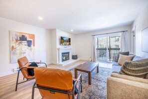 A modern living room with a fireplace, a sofa, a coffee table, and a view of the outdoors through large windows.