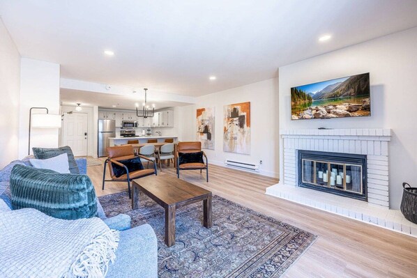 A modern living room with a fireplace, a dining area, and a kitchen in the background.