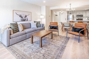 A modern living room with a sofa, coffee table, and two chairs. A kitchen is visible in the background.