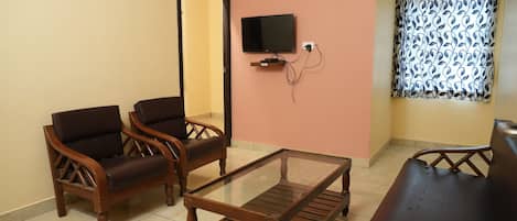 A room with two wooden chairs, a glass-top coffee table, a sofa, a TV mounted on the wall, and a window with patterned curtains.