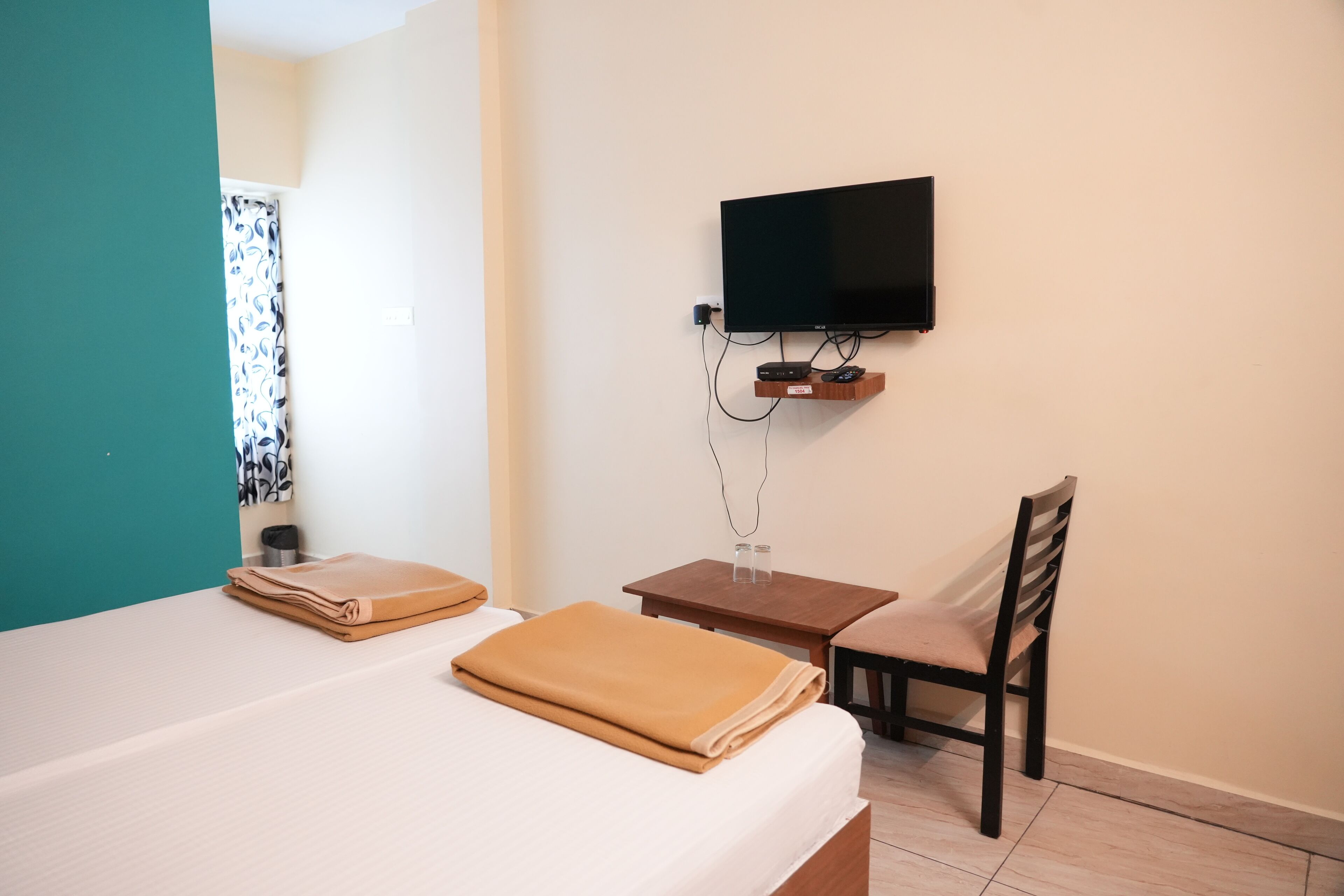 A hotel room with a bed, a TV mounted on the wall, a small wooden table, a chair, and a bathroom with a shower curtain.