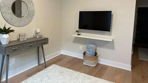 Una habitación con un escritorio de color gris, un espejo redondo, un televisor de pantalla plana montado en un estante blanco, una pequeña planta en maceta y una alfombra blanca sobre el piso de madera.