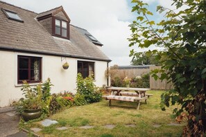 A white cottage with a sloped roof, a wooden fence, and a picnic table in the yard.
