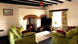 A living room with green upholstered sofas, a fireplace, a table, and a window with curtains. - Cosy Stone Cottage w/ Log Burner & Views (Nolton)