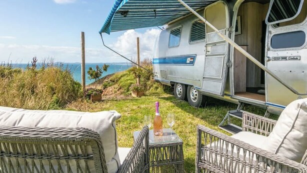 A camper van with a striped awning, a table with a bottle and glass, and wicker chairs. - Airstream with Stunning Coastal Views. (Nolton Haven)
