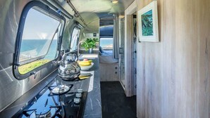 A compact kitchen with a stove, kettle, and a window overlooking the sea. - Airstream with Stunning Coastal Views. (Nolton Haven)