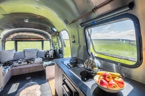 Interior de autocaravana compacta y moderna, con comedor, zona de estar con sofá y cojines, y un ventanal con vistas a un campo verde.