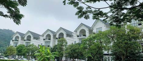 A row of white buildings with arched windows and balconies, surrounded by green grass and trees.