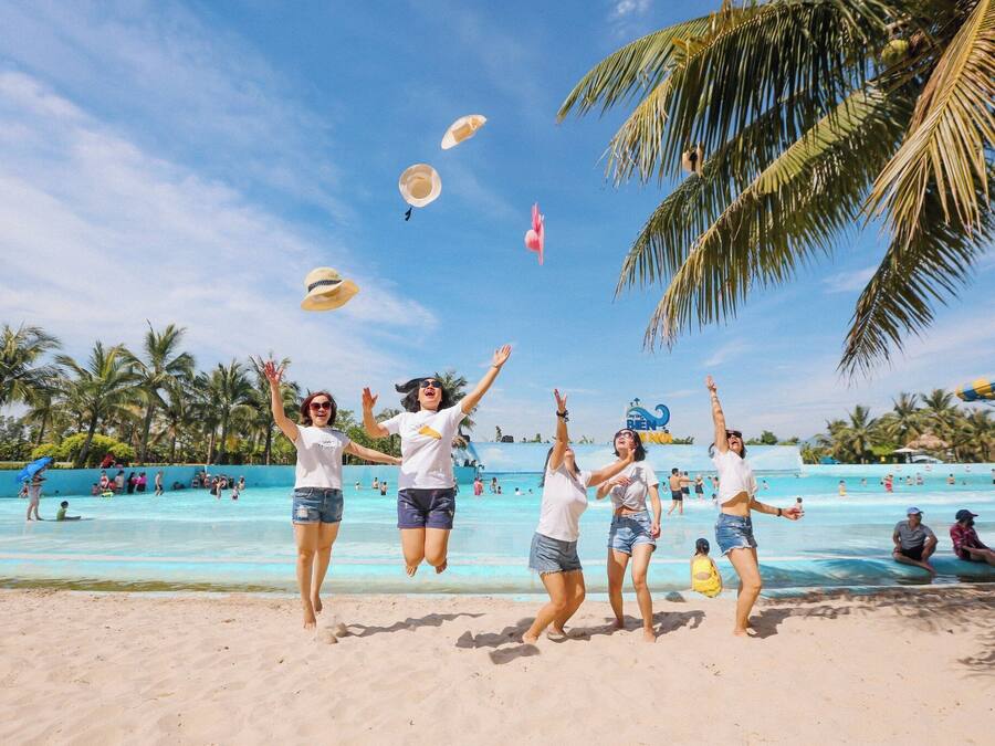 A beach scene with people jumping and playing in the sand, palm trees, and a water park in the background.