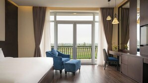 Una habitación de hotel con una cama grande, un escritorio con una silla, vistas a un paisaje verde a través de un ventanal amplio y un balcón con cortinas.